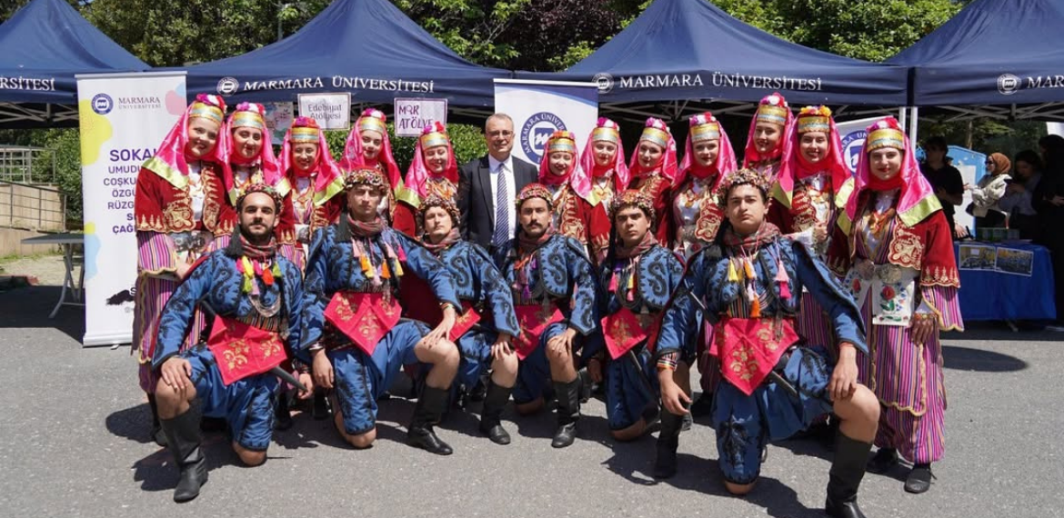 Marmara Üniversitesi Folklor Kulübü üyeleri sahnede yan yana dizilmiş, geleneksel kıyafetleri içinde, yüzlerinde büyük bir gülümsemeyle el ele tutuşarak coşkulu bir şekilde dans ediyorlar.